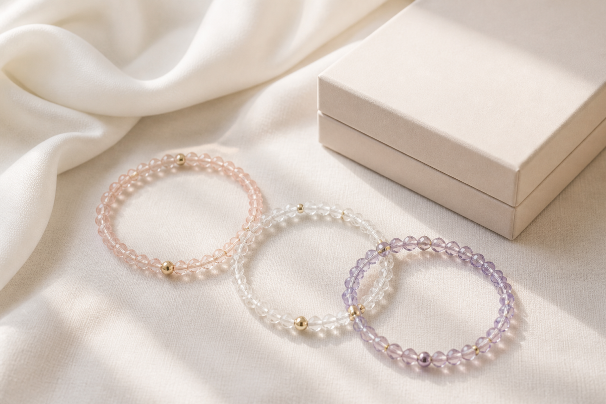 Three simple crystal bead bracelets on pale fabric beside a minimal jewelry box