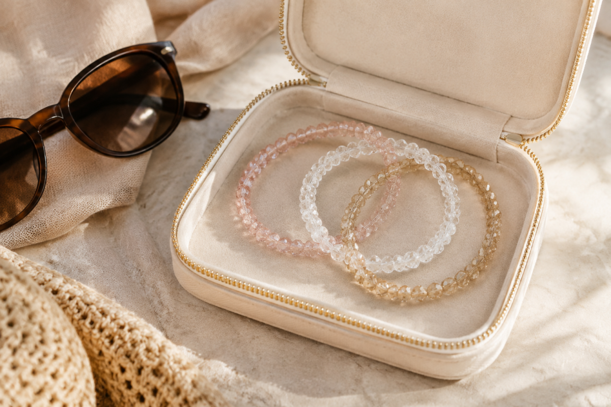 Three crystal bead bracelets in a cream travel pouch beside neutral resort fabric and sunglasses
